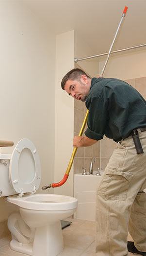 Man in khakis and a work shirt using a rod to unclog a toilet